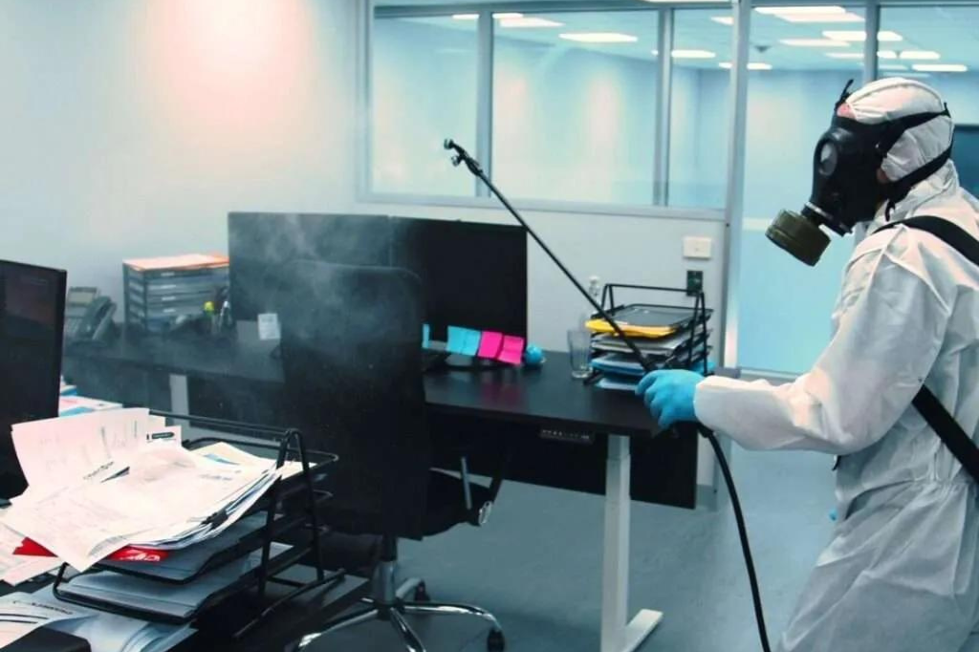 Person in hazmat suit with gas mask disinfecting an office desk, surrounded by office equipment and papers.