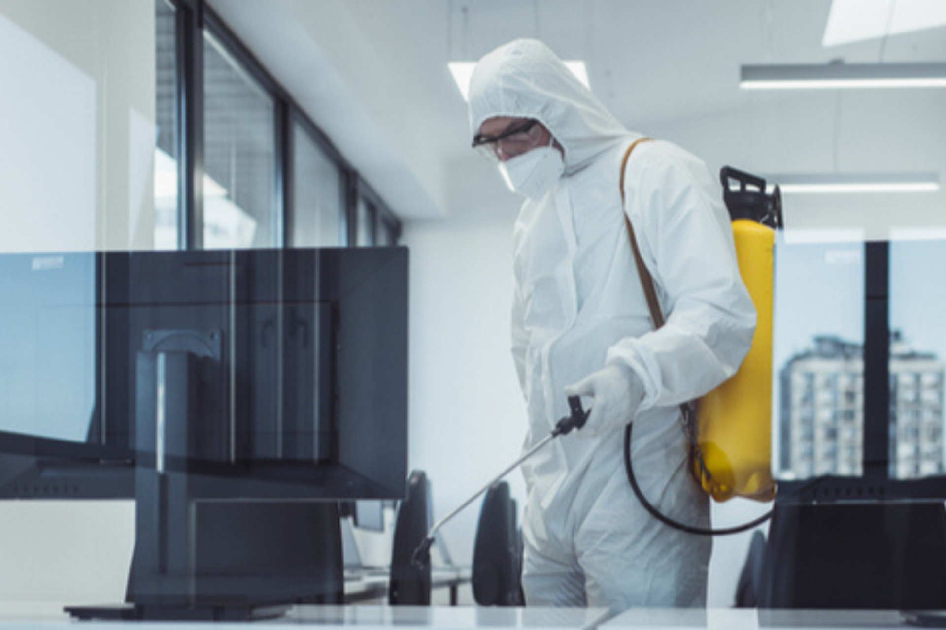 A person in a protective suit and mask spraying an office space with a spray device.