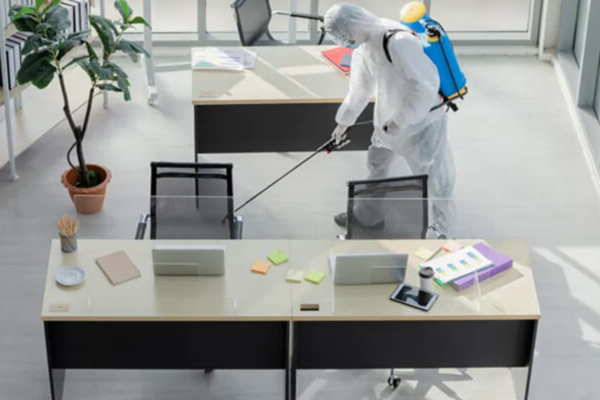 Person in protective gear disinfecting an office desk with a spray device.