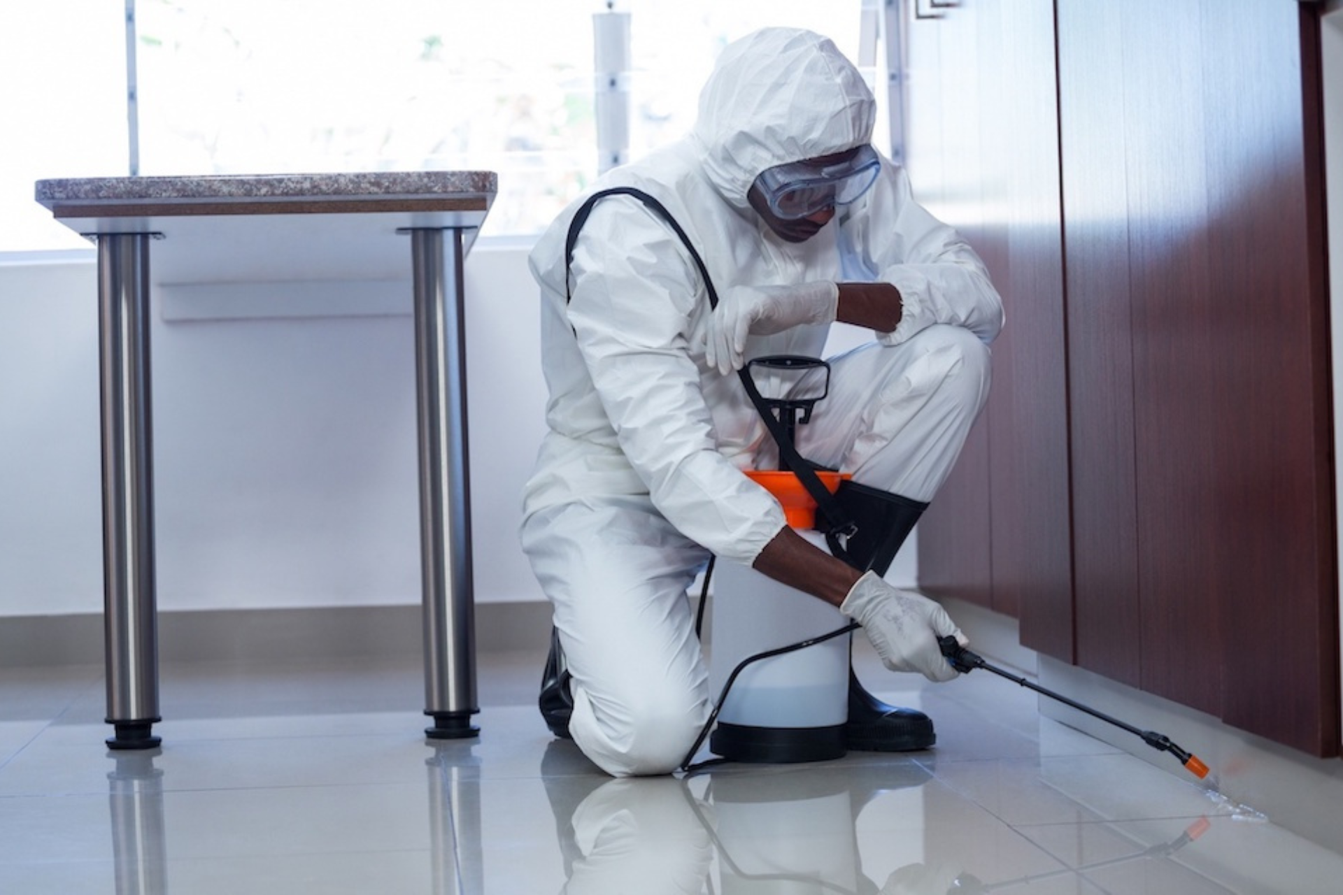 A pest control worker in a protective suit and mask sprays chemicals near a kitchen cabinet inside a home.