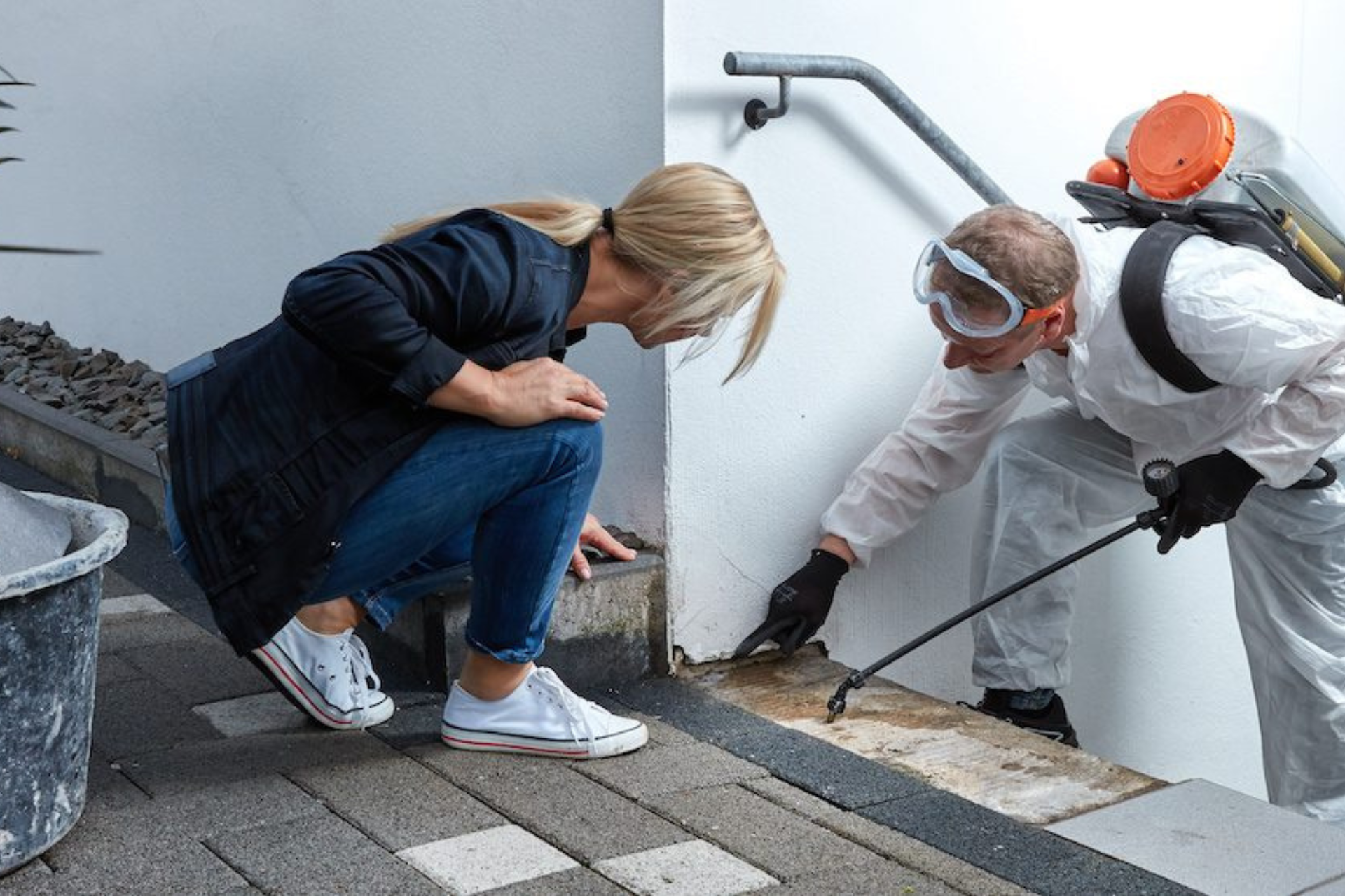 A woman observes a pest worker in protective gear using equipment to inspect or clean a building's exterior corner.