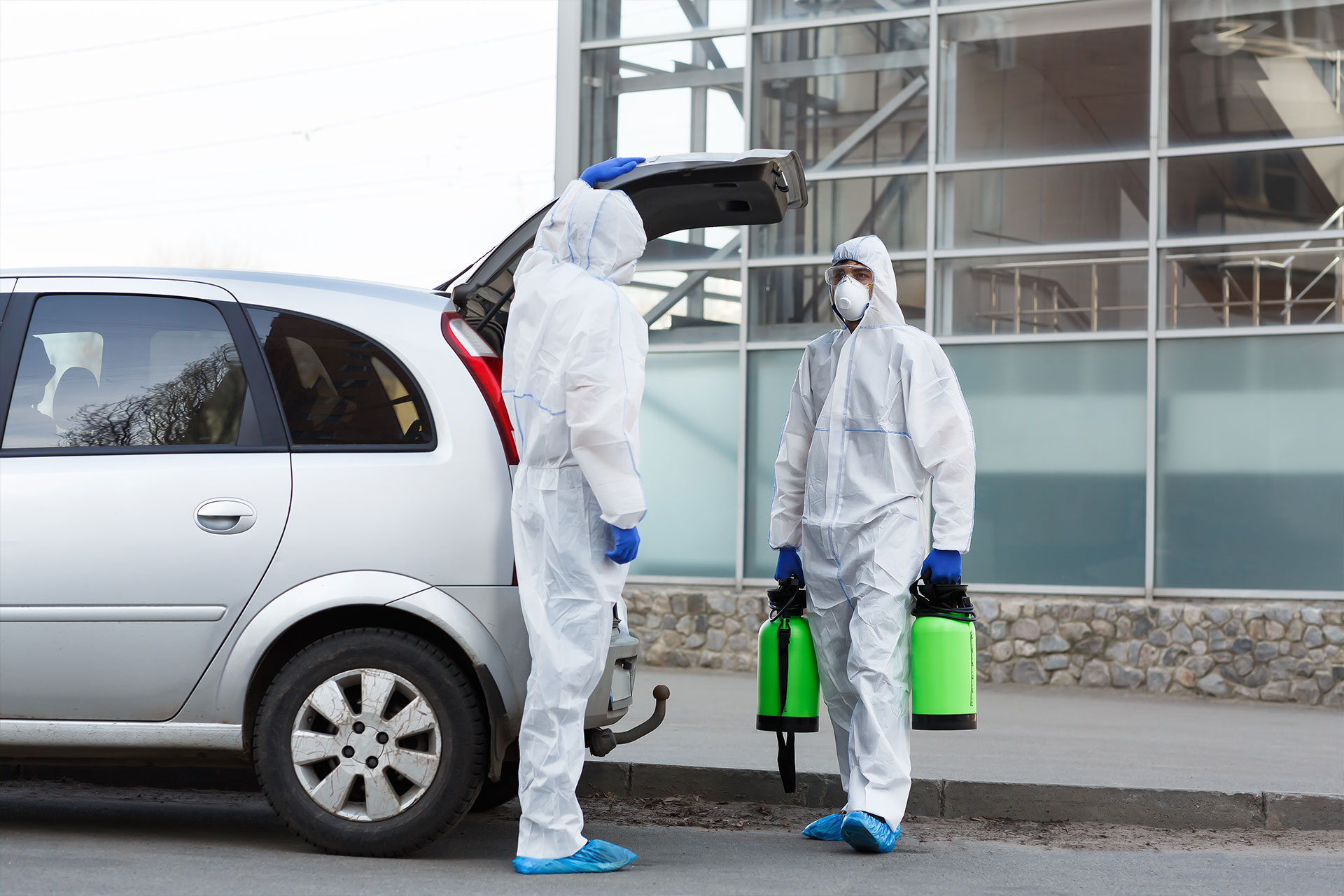 Two pest worker in hazmat suits holding disinfectant sprayers beside a car with an open trunk, in an urban setting.