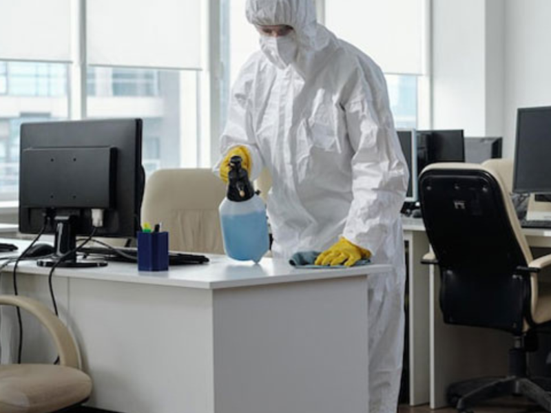 Person in protective gear disinfecting an office desk and computer with a spray bottle.