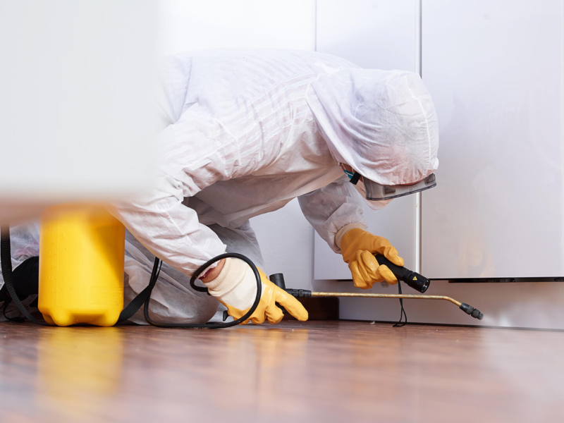 Pest control worker in protective suit using equipment to inspect underneath kitchen cabinets.